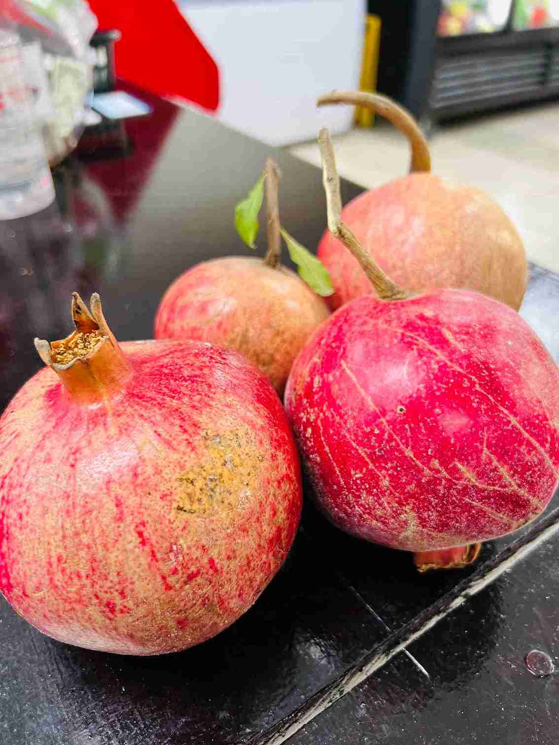 Four fresh, red pomegranates with stems and small green leaves on a display counter at Bismillah Grocery & Meat Market.